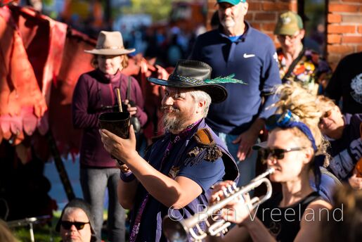 Man in a feathered wide-brimmed fedora playing cowbell at 'HONK!' with a knowing look in his eyes